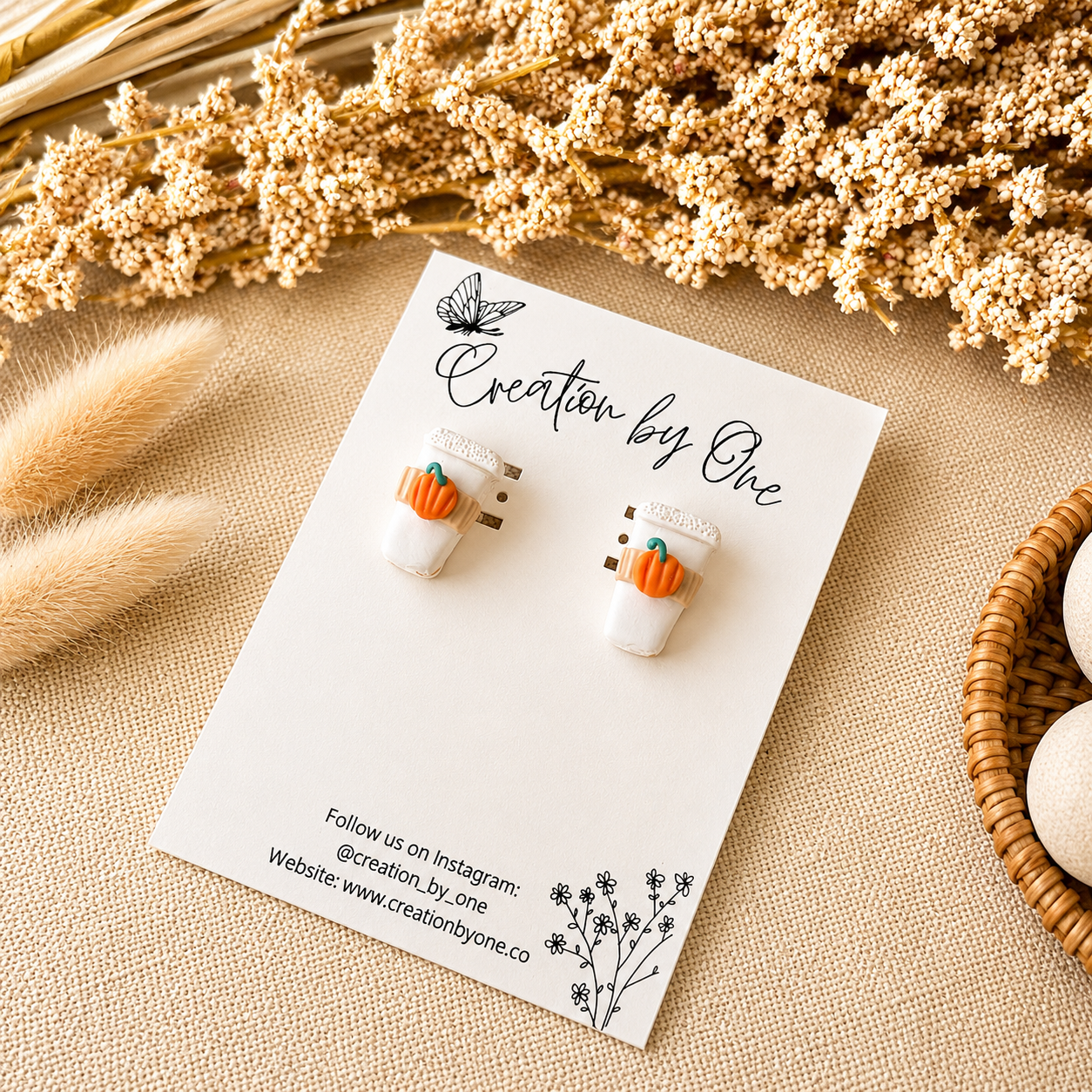 Pair of earrings on a card with 'Creation by One' branding, surrounded by dried flowers and a woven basket.