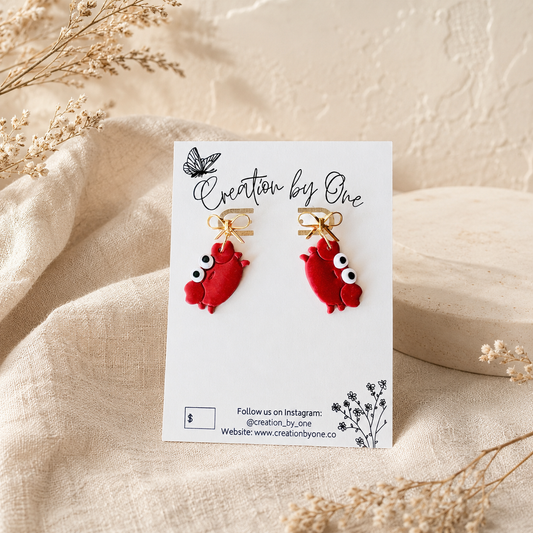 A pair of red fish-shaped earrings with gold fishhook wires and white eyes, displayed on a white card labeled 'Creation by One' on a beige fabric background with dried flowers.