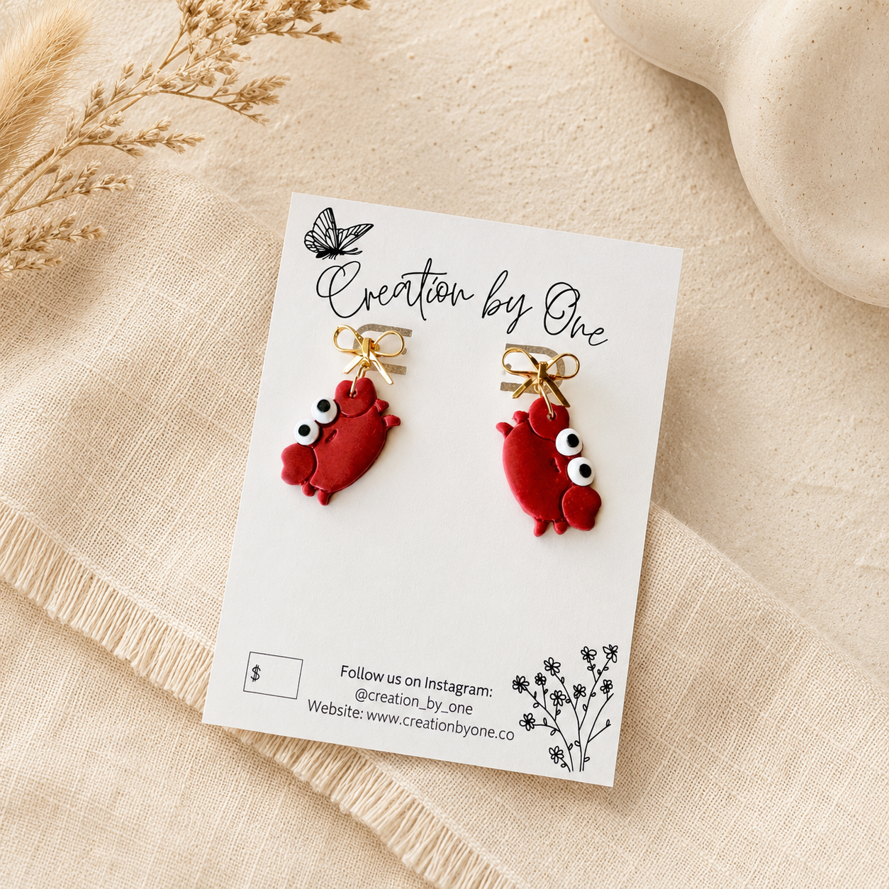 A pair of red cartoon crab earrings with large white eyes and gold posts, displayed on a white card labeled 'Creation by One' on a beige fabric background.