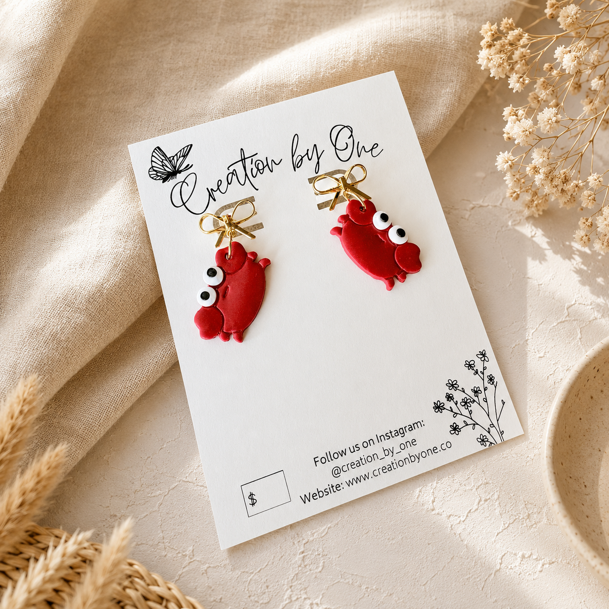 A pair of red lobster-shaped earrings with white eyes and gold loops, displayed on a white card labeled 'Creation by One' on a beige fabric background with dried flowers.