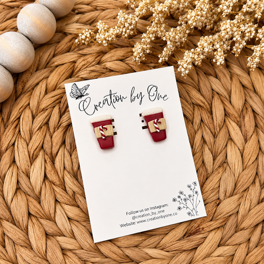 Handmade red holiday coffee cup stud earrings with candy cane details displayed on a white jewelry card against a warm woven boho background with natural wooden beads and dried florals. Perfect festive Christmas accessory for everyday holiday wear.