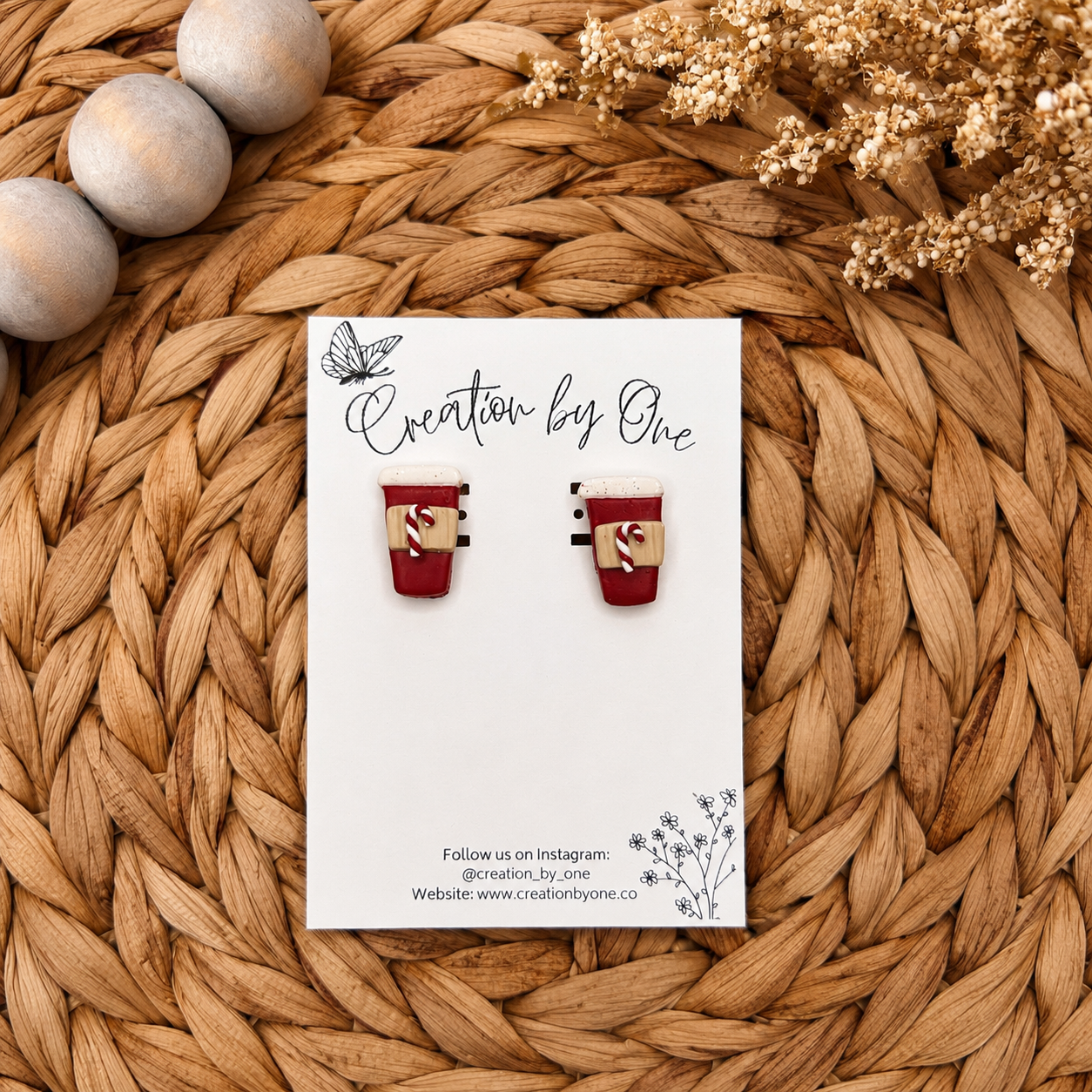 Handmade red holiday coffee cup stud earrings with candy cane details displayed on a white jewelry card against a warm woven boho background with natural wooden beads and dried florals. Perfect festive Christmas accessory for everyday holiday wear.