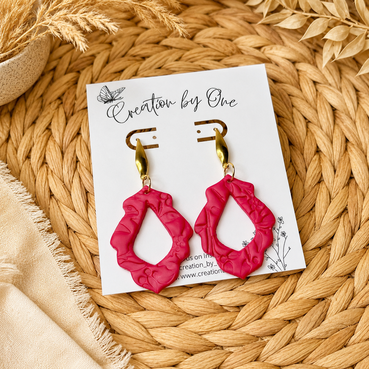 Pink earrings on a white card with 'Creation by One' branding, placed on a woven surface.