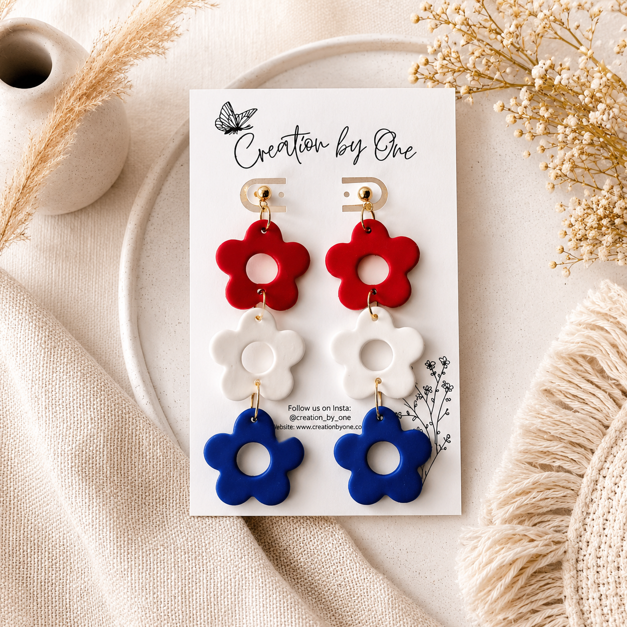Set of red, white, and blue flower-shaped earrings on a card labeled 'Creation by One' with decorative elements.