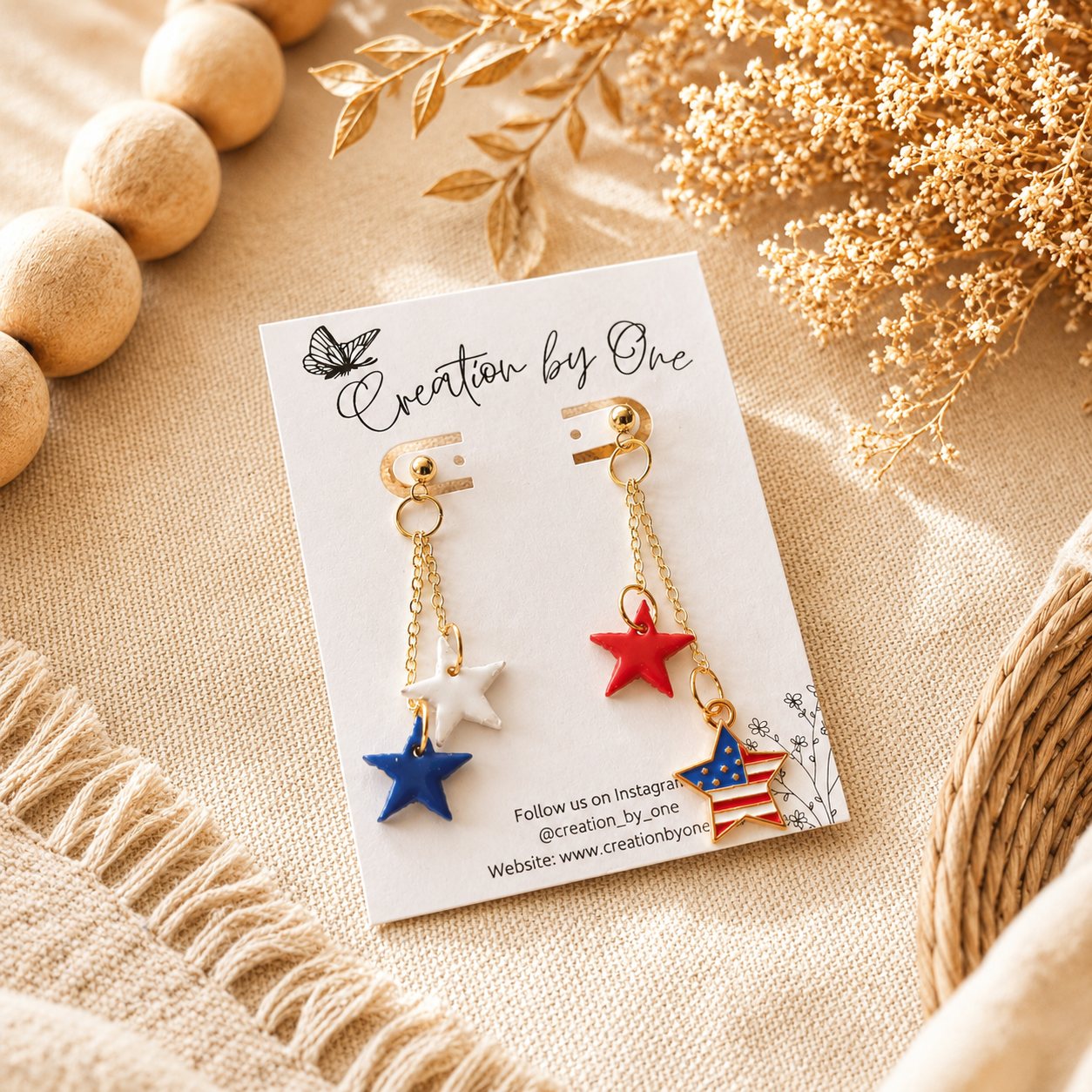 Pair of earrings with star and American flag design on a 'Creation by One' branded card, surrounded by decorative elements.