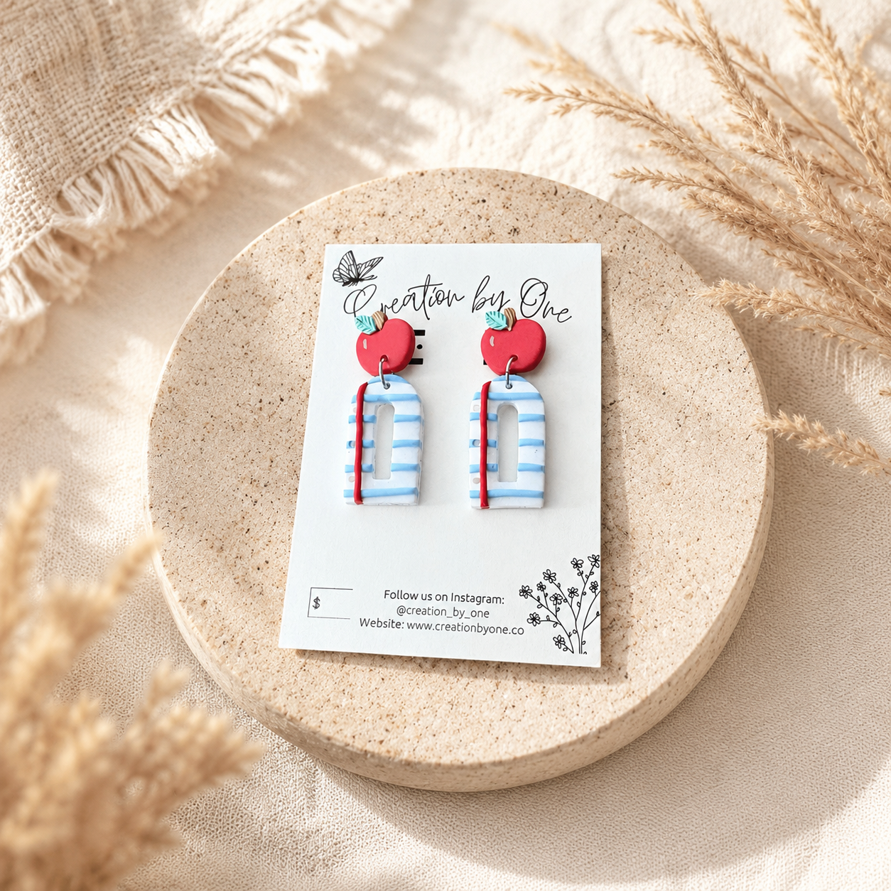 Handmade polymer clay teacher earrings displayed on a Creation by One jewelry card featuring bright red apple studs paired with blue-and-white striped notebook-style dangles accented with a red pencil detail. The card is styled on a soft neutral stone tray with warm beige linen fabric and dried pampas grass, creating a clean, modern boho product display with natural lighting.