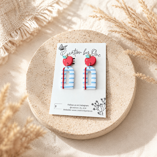 Handmade polymer clay teacher earrings displayed on a Creation by One jewelry card featuring bright red apple studs paired with blue-and-white striped notebook-style dangles accented with a red pencil detail. The card is styled on a soft neutral stone tray with warm beige linen fabric and dried pampas grass, creating a clean, modern boho product display with natural lighting.