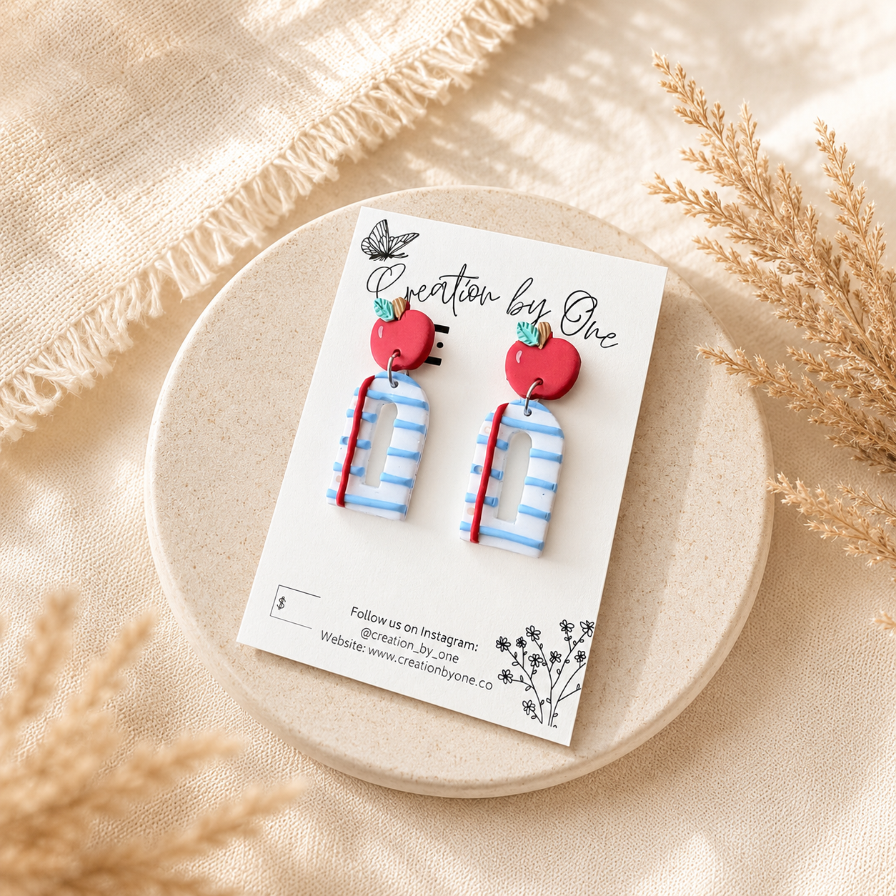 Handmade polymer clay teacher earrings displayed on a Creation by One jewelry card featuring bright red apple studs paired with blue-and-white striped notebook-style dangles accented with a red pencil detail. The card is styled on a soft neutral stone tray with warm beige linen fabric and dried pampas grass, creating a clean, modern boho product display with natural lighting.
