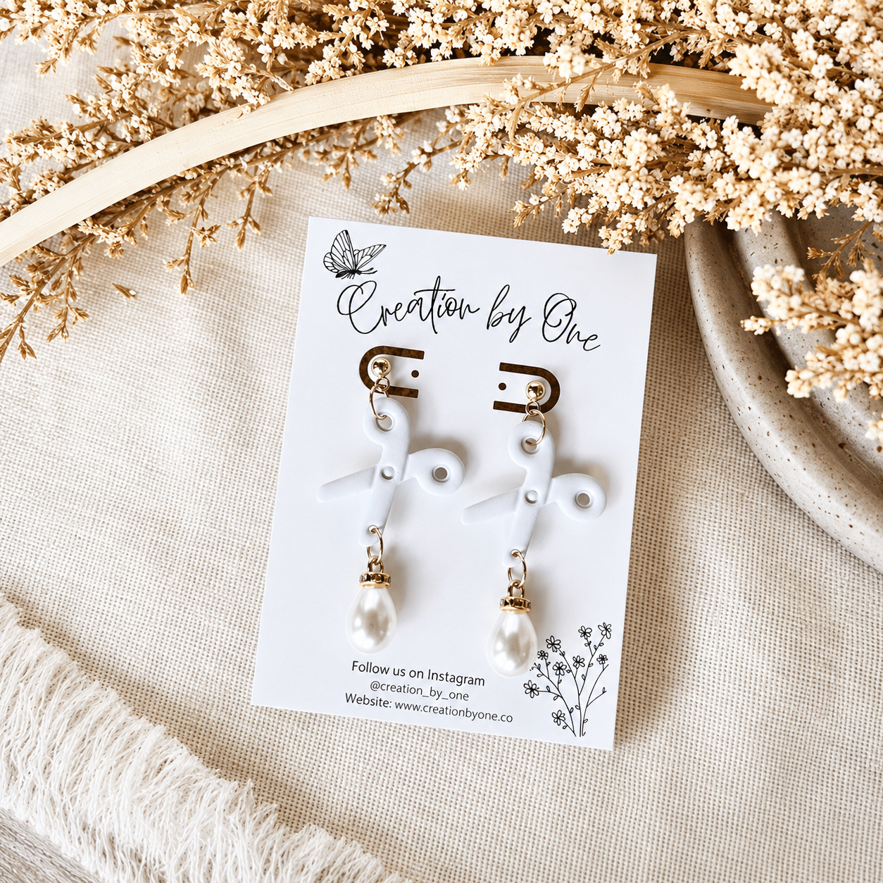 Pair of earrings on a 'Creation by One' branded card with dried flowers in the background.