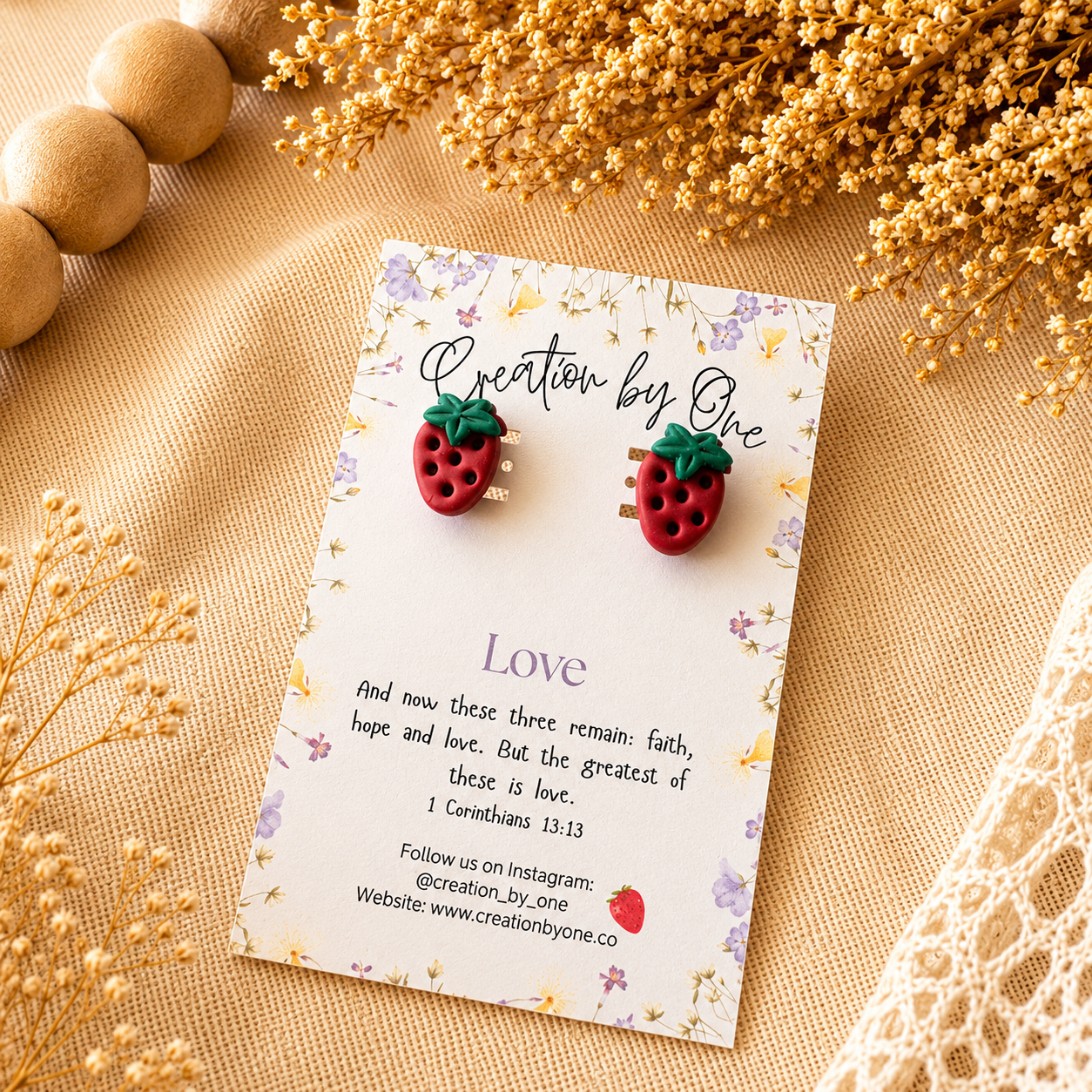 Strawberry-shaped earrings on a 'Creation by One' branded card with decorative elements on a beige surface.