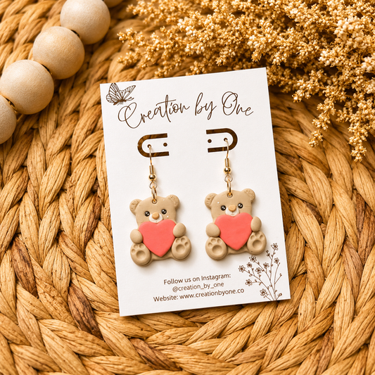 Teddy bear earrings on a 'Creation by One' branded card with dried flowers and beads in the background.