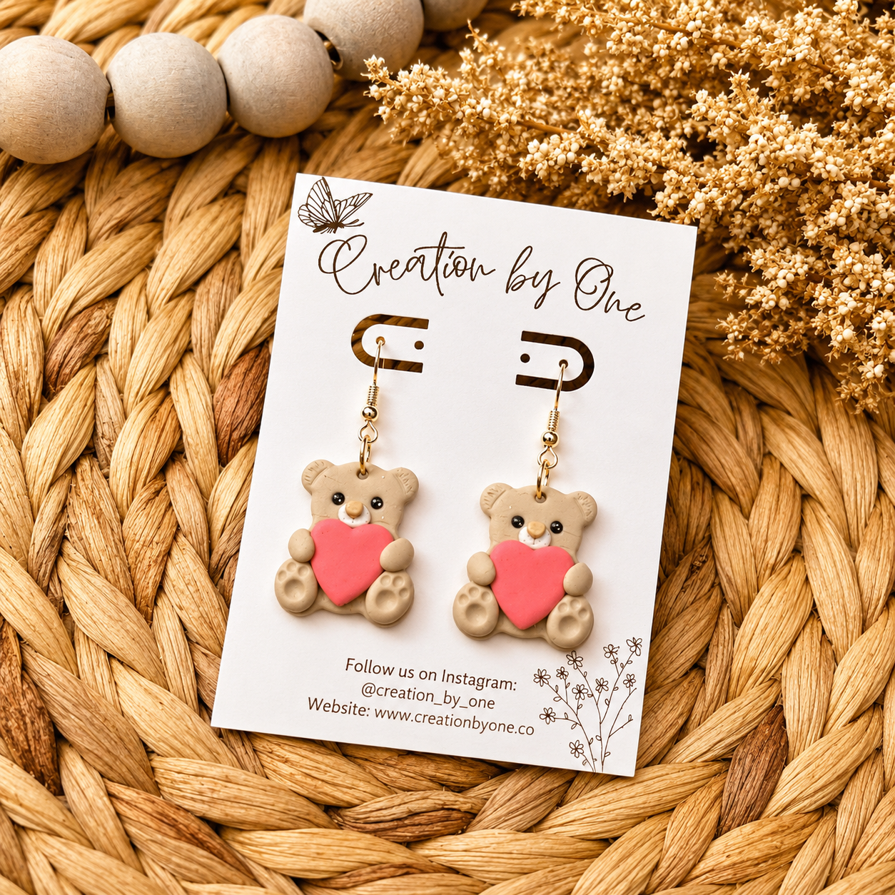 Teddy bear earrings with a heart on a 'Creation by One' branded card, placed on a woven surface.