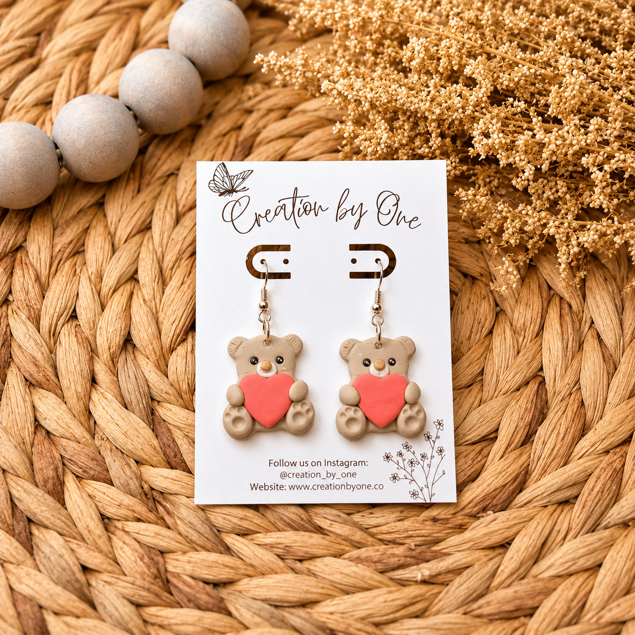 Teddy bear earrings with a heart on a branded card, placed on a woven surface.