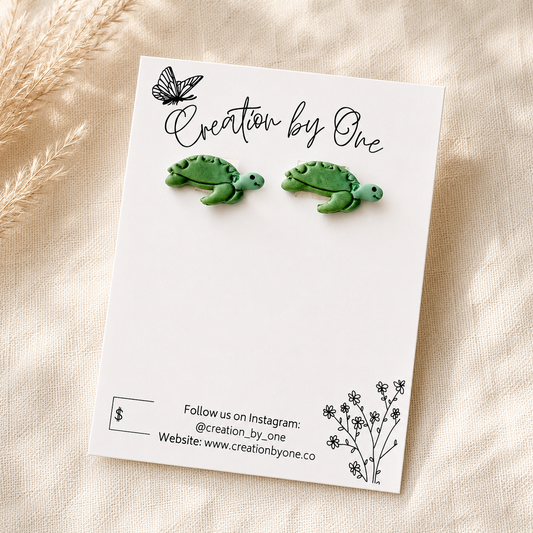 A pair of green turtle stud earrings displayed on a white card that reads 'Creation by One,' photographed on a soft beige fabric background with dried grass as styling props.