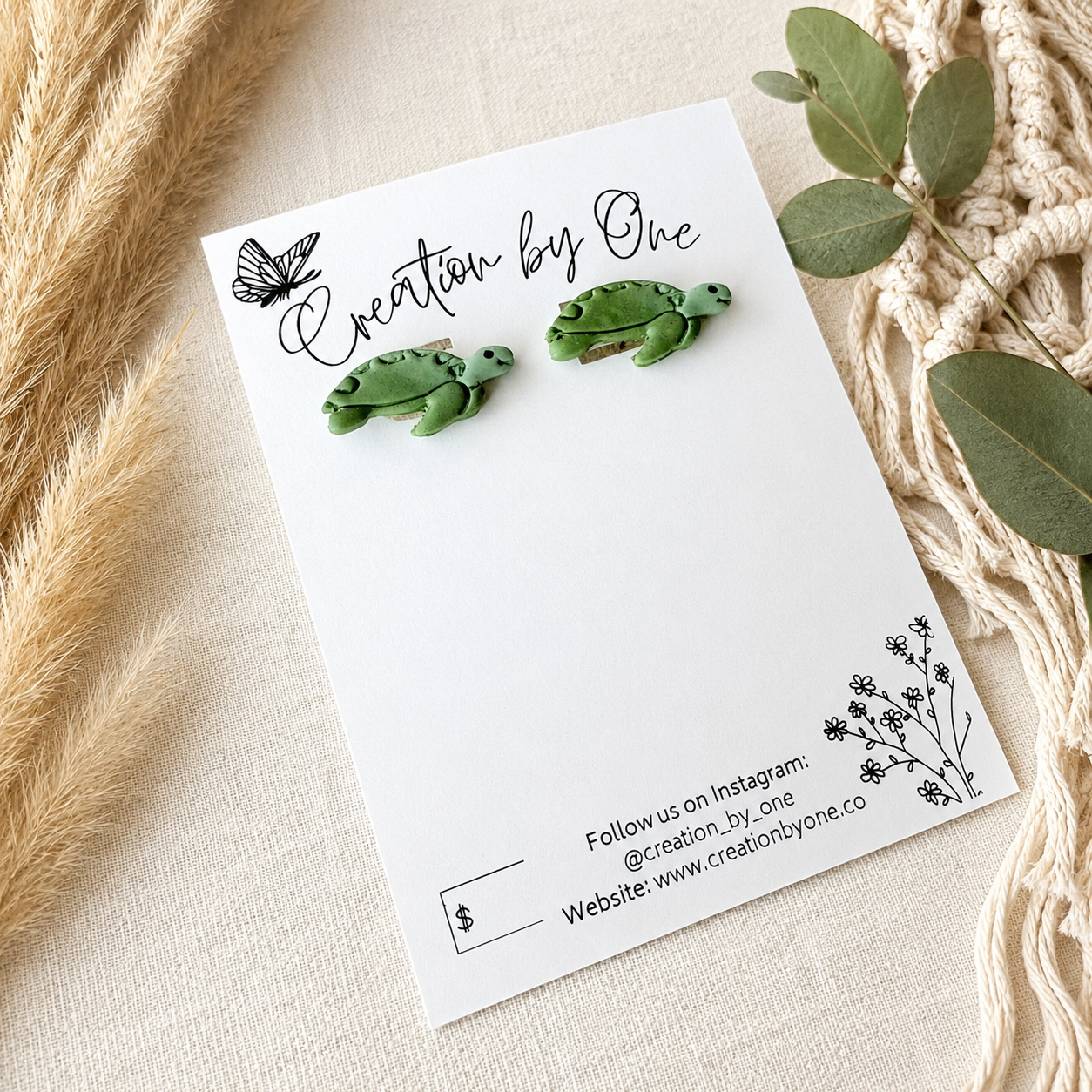 A pair of small green turtle stud earrings displayed on a white branded card, arranged on a neutral fabric surface with dried grass, green leaves, and a macrame textile around it.