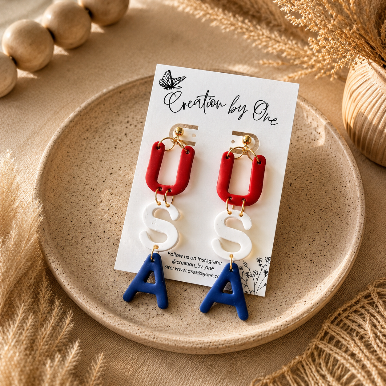 Earrings with red and blue letters on a 'Creation by One' branded card, placed on a beige plate.