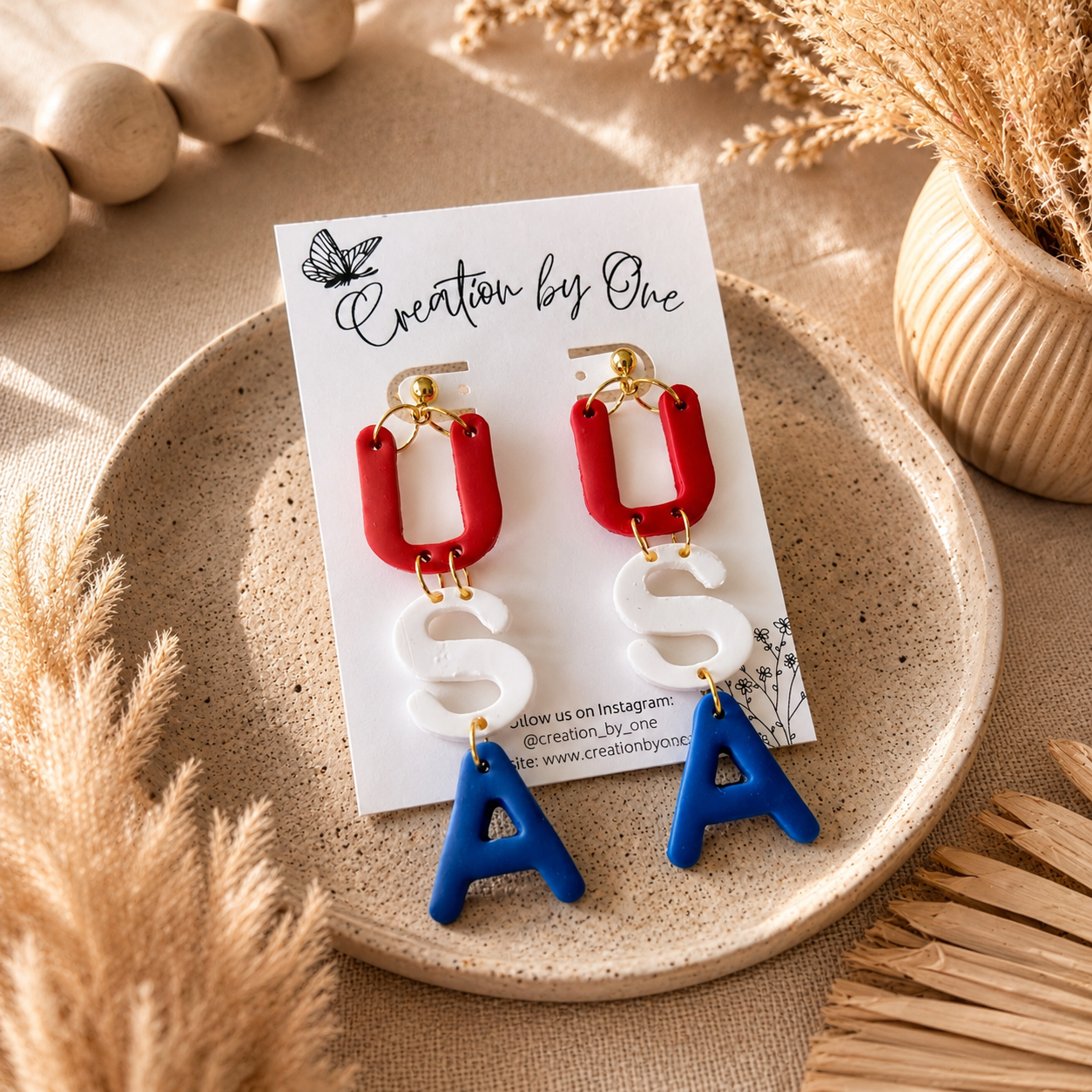 Pair of earrings on a card with 'Creation by One' branding, displayed on a textured surface with decorative elements.