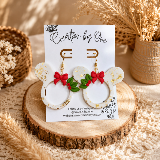 Handmade white Christmas mouse-shaped dangle earrings with gold leaf flakes, red bows, and holly accents displayed on a jewelry card against a soft neutral boho fabric background with dried florals.