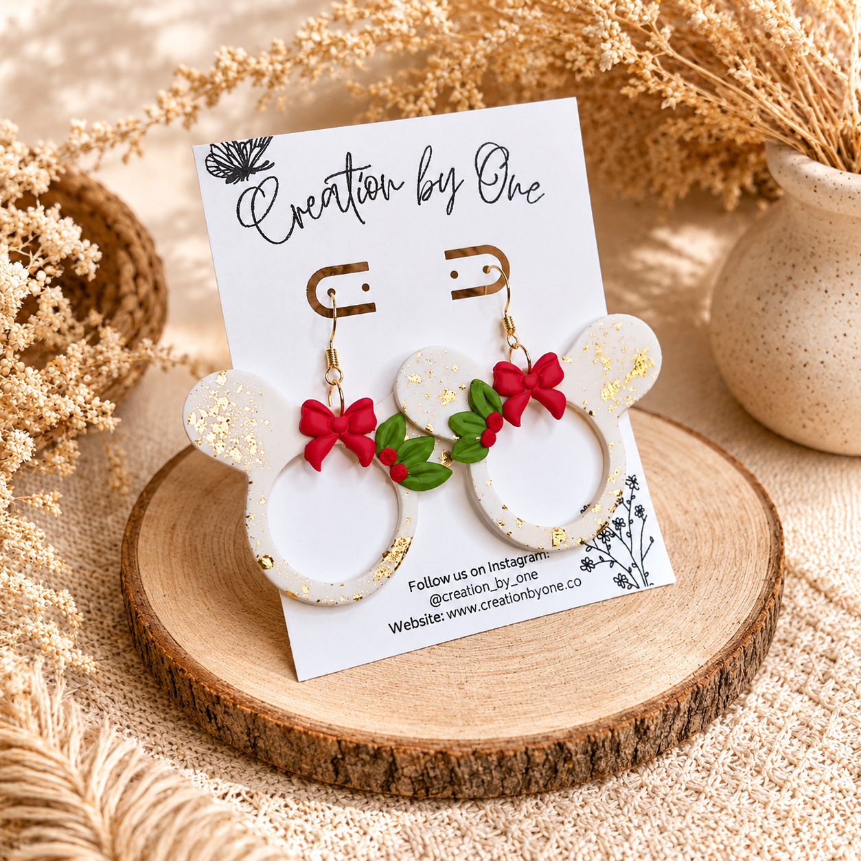 Handmade white Christmas mouse-shaped dangle earrings with gold leaf flakes, red bows, and holly accents displayed on a jewelry card against a soft neutral boho fabric background with dried florals.