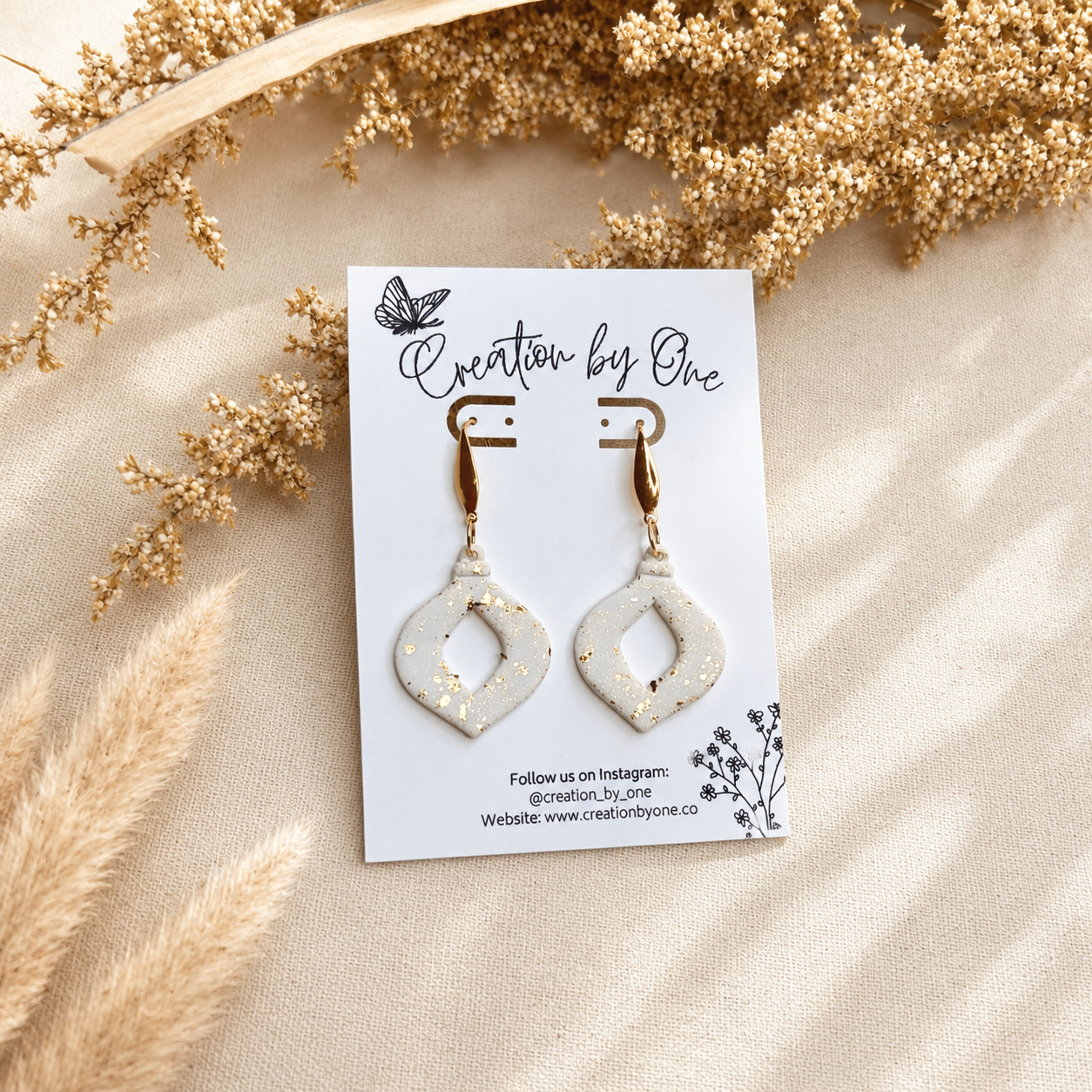 Handmade white polymer clay geometric dangle earrings with gold leaf speckles and gold-tone hooks displayed on a Creation by One jewelry card against a neutral linen background with dried boho florals.