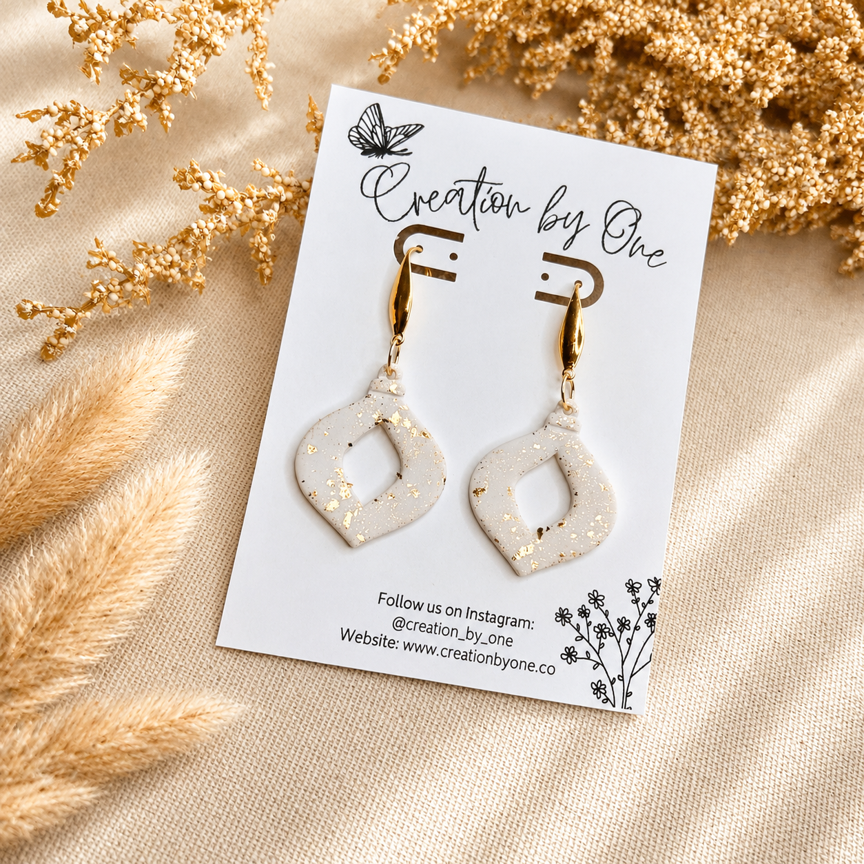 Handmade white polymer clay geometric dangle earrings with gold leaf speckles and gold-tone hooks displayed on a Creation by One jewelry card against a neutral linen background with dried boho florals.