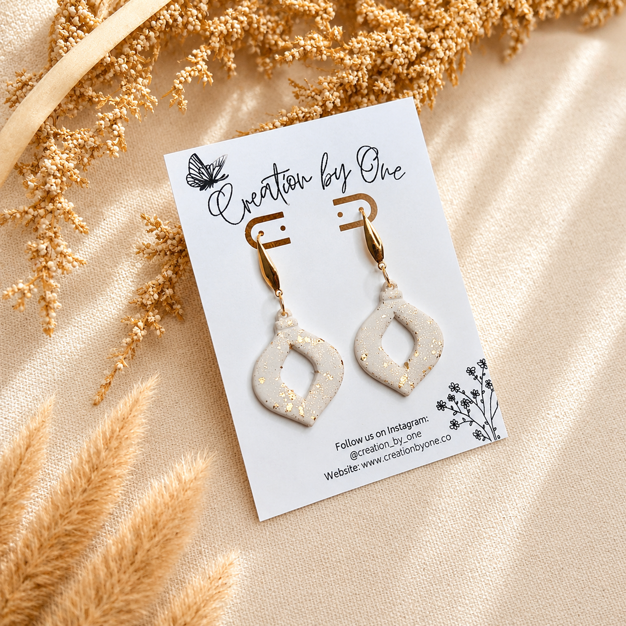 Handmade white polymer clay geometric dangle earrings with gold leaf speckles and gold-tone hooks displayed on a Creation by One jewelry card against a neutral linen background with dried boho florals.