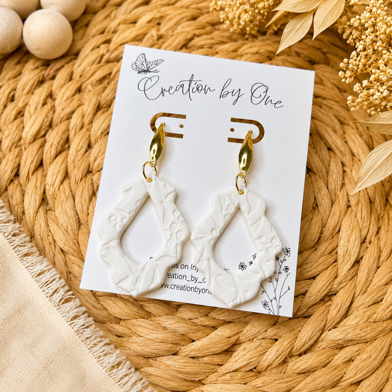 White earrings with gold accents on a 'Creation by One' branded card, placed on a woven mat.