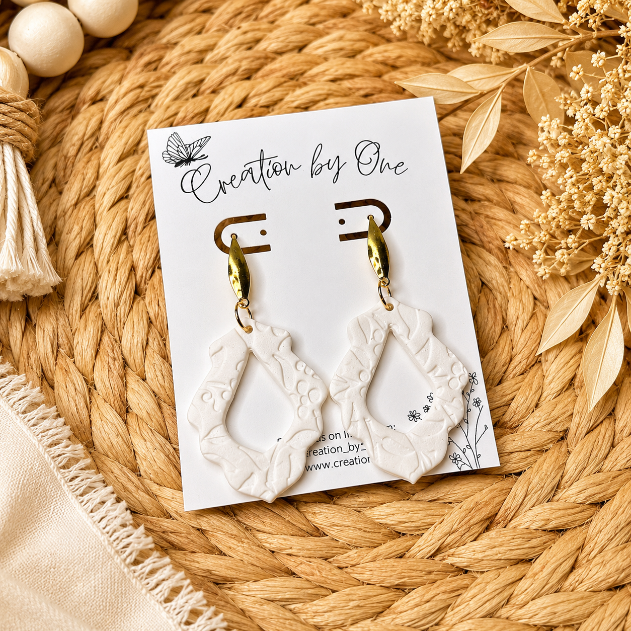 White earrings on a 'Creation by One' branded card with decorative elements on a woven surface.