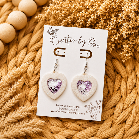 Heart-shaped earrings on a card with 'Creation by One' branding, placed on a woven surface.