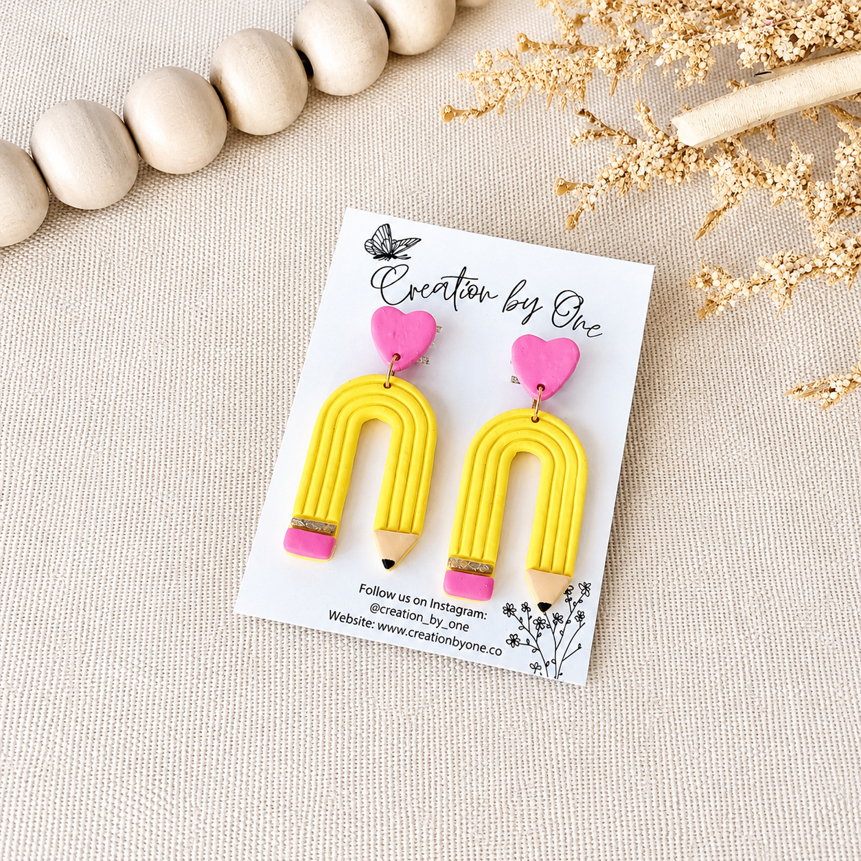 Yellow heart-shaped earrings on a card with 'Creation by One' branding, placed on a textured surface.
