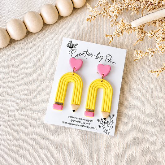 Yellow arch-shaped earrings with pink hearts on a 'Creation by One' branded card, surrounded by wooden beads and dried flowers.