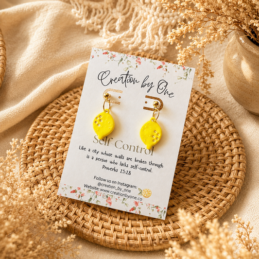 Yellow earrings on a 'Creation by One' branded card with decorative elements on a textured surface.